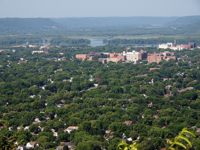 [Amid the slightly open spaces between trees are glimpses of homes. In the further distance are the multi-storied buildings in town. Further in the background is the river and the bluffs on the Minnesota side of the river.]