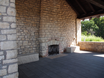[A light-colored stone fireplace set into a stone wall holding a wooden-beamed roof. The floor is flagstone.]
