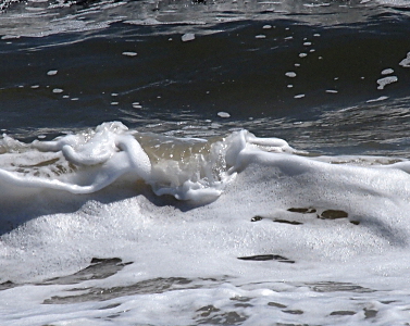 [The foreground is almost completely covered with teeny white bubbles. At the center of an image is a small section of water breaking over the bubbles and looks like a waterfall as the water is bubble-free. Behind this is still-looking dark blue water with a small scattering of white bubbles.]