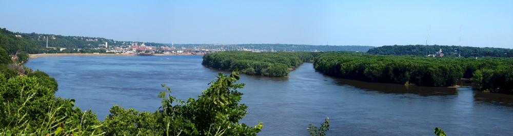 [A view of the river and islands in it from a vantage point just a bit higher than the river level. The many buildings of the city can be seen in the distant shore.]