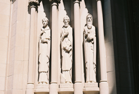 [Statues which appear to be carved into the building of three tall thin men with a column between each of them. The one on the far left is clean shaven, the middle one has a small beard and the one on the right seems to have a long beard. All three appear to be holding their clothing capes closed.]